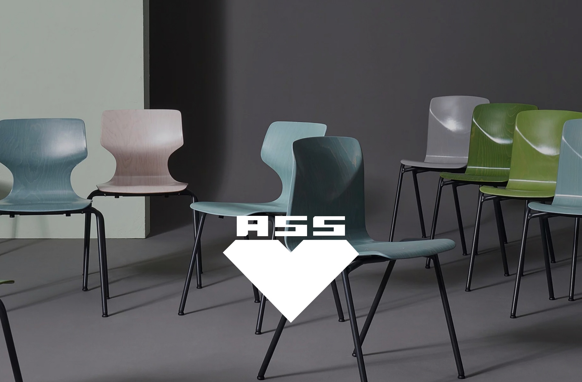 Eine Vielzahl verschiedenfarbiger Plastikstühle in Pastelltönen ist in einem Raum mit dunkelgrauer Wand aufgereiht. Die Stühle stehen paarweise nebeneinander und sind durch ein weißes Symbol mit den Buchstaben 'ASS' gekennzeichnet. 